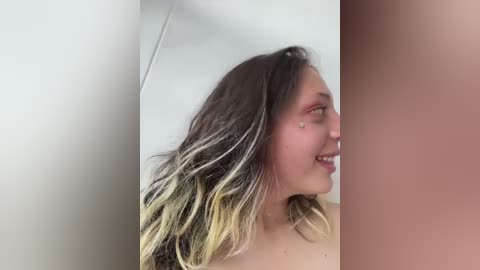 Video of a young woman with long, wavy hair featuring blonde and dark brown streaks. She has a light skin tone and is smiling. The background is a plain, light-colored wall.