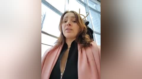 Video of a woman with light skin, shoulder-length brown hair, wearing a pink blazer over a black top, standing in front of a large window with a deer head mounted on the wall in the background.