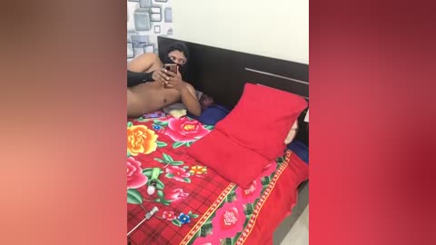 Video of a naked person with medium brown skin, long black hair, and a medium build, lying on a bed with a red and floral blanket, holding a smartphone.