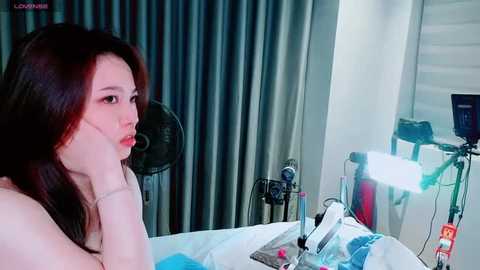 Video of a young Asian woman with fair skin, long black hair, and a neutral expression, seated in a modern room with grey curtains and studio lighting, holding a camera.