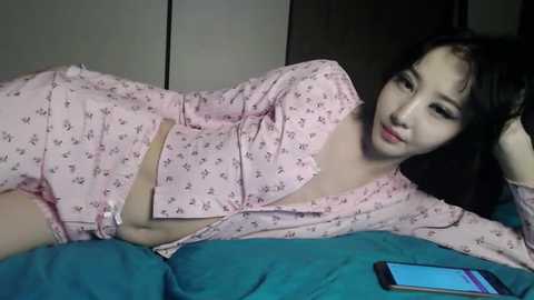 A video of an Asian woman with fair skin, lying on a teal bedspread, wearing a pink floral nightgown, revealing her breast, in a dimly lit bedroom.