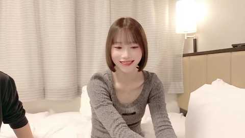 Video of a young Asian woman with shoulder-length brown hair, smiling, wearing a gray ribbed long-sleeve top, sitting on a bed with white sheets, in a modern hotel room with beige curtains and a bedside lamp.