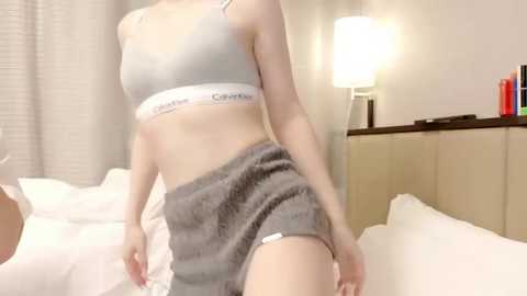 Video of a fair-skinned woman in a Calvin Klein sports bra and grey shorts, posing on a white bed with beige curtains and a lit lamp in the background.