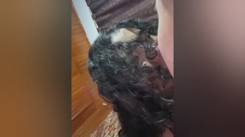 Video of a man with long, dark, curly hair, lying on a wooden floor, partially obscured by a brown curtain.