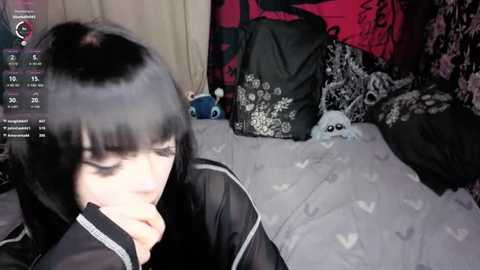 Video of a young woman with black hair, pale skin, and black clothing, lying on a bed with a black and white patterned blanket. Background shows a pink and black patterned wall, stuffed animals, and a black bag.
