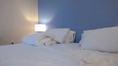 A minimalist bedroom with a white bed, blue wall, and a bedside table with a white lamp. The bed has neatly folded white sheets and pillows, creating a clean and serene atmosphere.