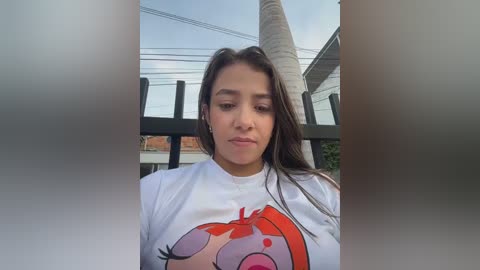 A video of a young woman with long brown hair, wearing a white t-shirt with a colorful cartoon apple graphic, sitting on a black metal fence in a suburban setting.