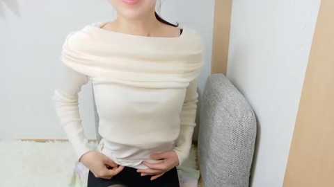 Video of a light-skinned woman with a slim build, wearing a long-sleeved, off-shoulder cream top and black pants, standing against a beige wall. She has light brown hair pulled back.
