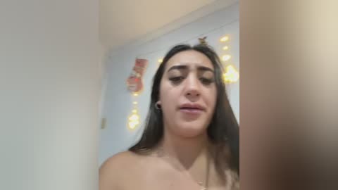 Media: Video of a young woman with long dark hair, fair skin, and full lips, standing topless in a dimly lit room with white walls and warm, yellow string lights.