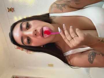 Video of a young woman with long dark hair, fair skin, and tattoos, licking a red lollipop, wearing a white top, in a brightly lit room.