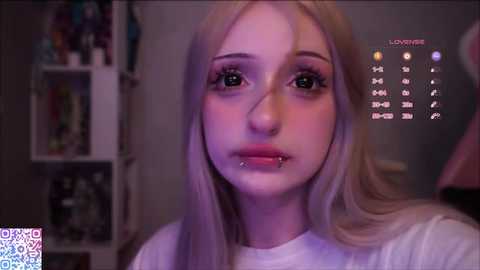 Video of a young woman with fair skin, long blonde hair, and multiple facial piercings, including a septum ring. She wears a white t-shirt. Background shows a dimly lit room with shelves and a wall calendar.