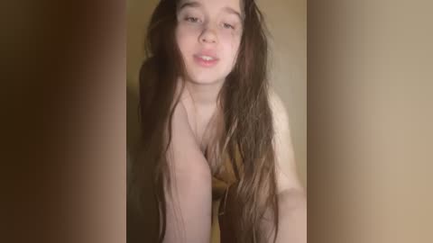 Video of a young, light-skinned woman with long, wet brown hair, wearing a brown bra, leaning forward, partially obscured by two vertical brown columns, creating a voyeuristic, intimate atmosphere.