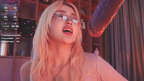 Video of a young, fair-skinned blonde woman with long hair, wearing glasses, a textured sweater, and a necklace, in a dimly lit room with a digital clock and blurred background.