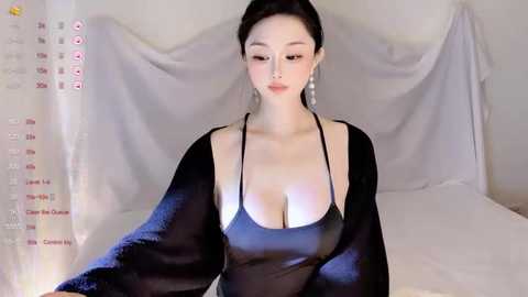 Video of an East Asian woman with fair skin, dark hair in a bun, wearing a sheer black top revealing cleavage, sitting on a bed with white sheets, draped in a beige cloth.