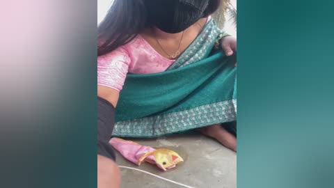 A video shows a woman in a pink, floral-patterned top and green sari, sitting on the ground. She holds a small, yellow snake with black markings, wearing a black mask.