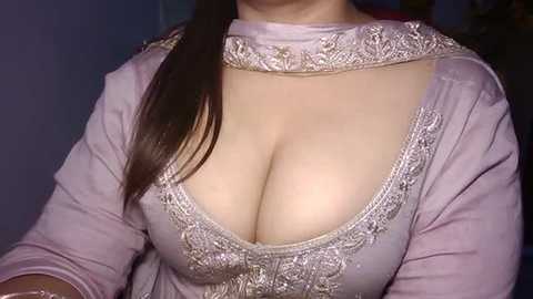 A video of a fair-skinned woman with long brown hair wearing a low-cut, pink dupatta adorned with intricate silver embroidery. The neckline reveals a significant cleavage. The background is dimly lit, with indistinct objects.