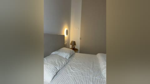 Video of a minimalist bedroom with a neatly made bed, two pillows, a wooden nightstand with a lamp, and a warm, ambient light.