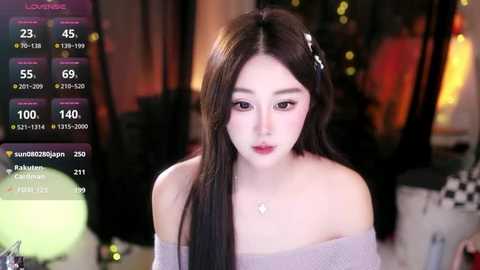Video of an East Asian woman with long black hair and pale skin, wearing a light off-shoulder top, set in a dimly-lit room with a cozy, bohemian ambiance.