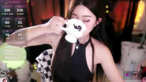 Video of a young East Asian woman with long black hair, wearing a black halter top, drinking from a white mug, in a dimly lit room with warm lighting and blurred background.