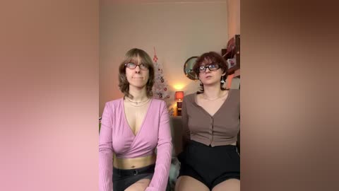 Video of two women sitting side by side in a dimly-lit room. One wears a pink cropped top and black shorts, the other a brown cardigan and black shorts. Both have glasses, and the room has a cozy, casual vibe.