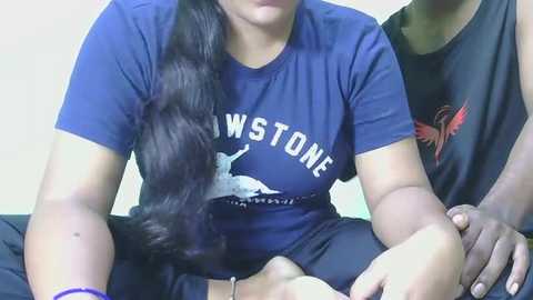 Video of a woman with long black hair wearing a blue \"Winston\" t-shirt, sitting with her legs crossed, and a black cat on her lap.