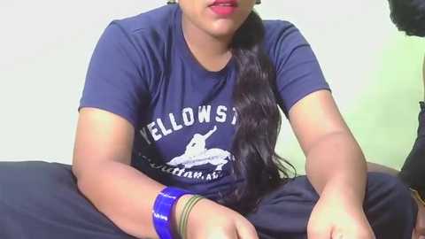 Video of a woman with medium brown skin, wearing a blue t-shirt with a logo, sitting on a black couch, with a blue bracelet on her wrist.