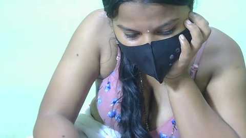 Video of a woman in a pink floral bra with black face mask, holding phone, in a simple room.