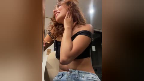 Video of a young woman with curly hair, wearing a black off-shoulder top and blue jeans, looking thoughtful while touching her neck. Background features a beige wall and a doorway.