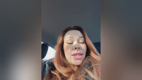 A video of a woman with long, wavy red hair, closed eyes, and slightly open mouth, seated in a car. The background is blurry, showing car interior elements.
