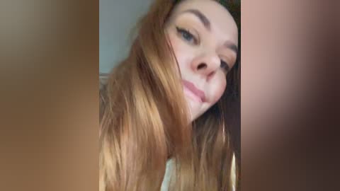 Video of a fair-skinned woman with long, wavy, light brown hair, wearing makeup, including dark eyeliner and mascara. Her expression is neutral. The background is blurred and neutral-toned.