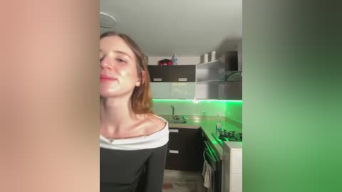 Video of a young woman with fair skin and brown hair, wearing a black off-the-shoulder top, standing in a modern kitchen with green LED lighting and dark cabinets.