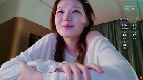 A candid video of an Asian woman with long brown hair and light skin, wearing a white off-shoulder sweater, smiling, in a dimly lit room with a digital overlay showing viewers and likes.