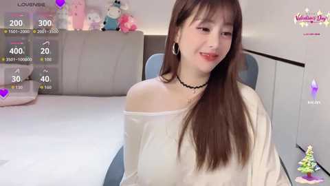 Video of an Asian woman with long brown hair and fair skin, wearing a white off-shoulder top and black choker. She sits on a bed in a bedroom with plush toys, pink decor, and a digital overlay showing likes and hearts.