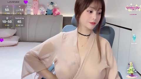 A young East Asian woman with fair skin, brown hair, and medium breasts, wearing a beige robe, sits in a chair. Background features a plush toy and a white wall.