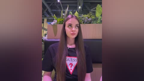 Media: Video of a young woman with long brown hair, fair skin, and green eyes, wearing a black \"Guess\" t-shirt, seated indoors with a lush green plant backdrop.