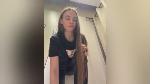 Video of a young woman with long, straight brown hair, wearing a black t-shirt with a \"St. Louis\" logo, in a dimly lit, beige-walled hallway.