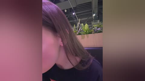 Video of a woman with long brown hair, partially cropped out, leaning over a plant in a modern, dimly lit room with exposed ceiling beams and greenery.