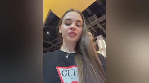 A video of a young woman with long, straight brown hair, wearing a black Gucci t-shirt, posing indoors with a playful expression.