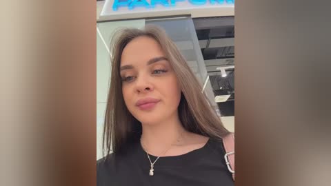 Video of a young Caucasian woman with long brown hair, fair skin, and a soft smile, wearing a black top, taken indoors in a dimly lit room with a blurred background.