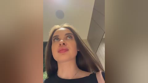 A video of a young woman with light skin, straight brown hair, and red lipstick, gazing upward, wearing a black top, in a modern indoor setting with beige walls and a lens flare.