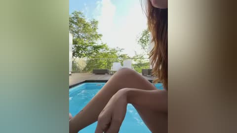 A video of a partially visible, nude woman with long, auburn hair, sitting by a blue pool surrounded by lush greenery and lounge chairs under a clear blue sky.