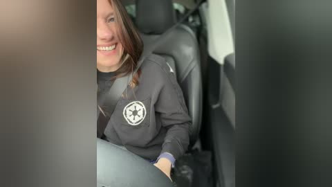 Video of a smiling woman in a gray jacket with a white police badge emblem, seated in a car, partially obscured by a black car door frame.