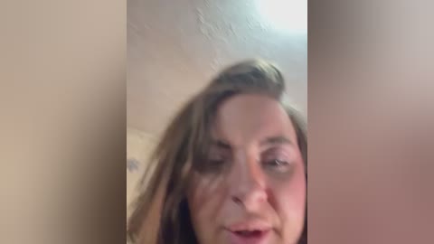 A blurred video of a middle-aged woman with fair skin, brown hair, and freckles, possibly from a bathroom, with a beige wall and a mirror in the background.