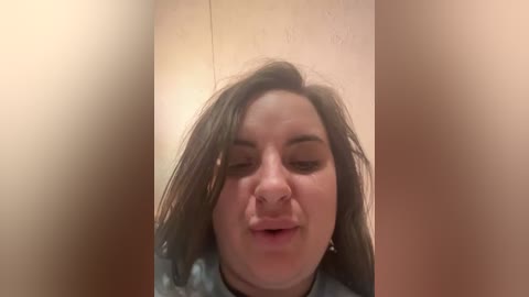 A video of a woman with fair skin, brown hair, and a prominent nose piercing, making a kissy face. She's wearing a dark shirt against a beige wall. The image is blurred, emphasizing the playful, candid nature.