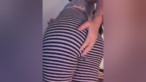 Video of a woman in tight, black-and-white striped leggings, showcasing her curvy, well-defined buttocks. Her hand rests on her right cheek, emphasizing the snug fit. Background shows a blurred indoor setting.