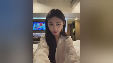 Media: A video of an Asian woman with long black hair, wearing a white jacket, sitting on a bed in a modern hotel room with a TV screen showing colorful images.