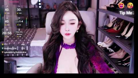 A video of an East Asian woman with long, wavy black hair, wearing a purple lace top and choker, seated in a modern living room with a gray couch and shelves filled with shoes.
