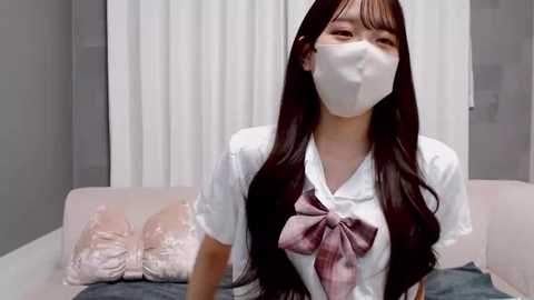 Video of an East Asian woman with long, straight black hair and fair skin, wearing a white mask, a white blouse with a pink bow, and a pink skirt, seated on a pink sofa with white cushions, in a minimalist room.