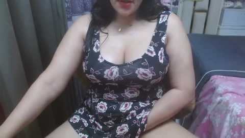 Video of a plus-sized woman with fair skin and dark hair, wearing a black floral dress, sitting on a couch.