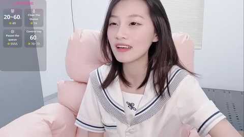 A video of an East Asian woman with straight black hair, fair skin, and a soft smile, wearing a white sailor-style dress, sitting on a pink chair in a minimalistic, light-colored room.